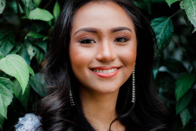 Smiling woman with flawless makeup and glowing skin, looking fresh and relaxed amidst green foliage