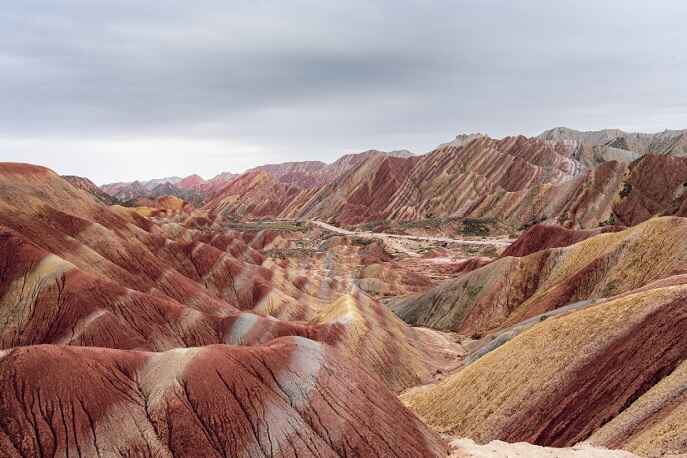 Colorful layered hills of Hormuz Island in Iran, showcasing vibrant red, orange, and yellow mineral-rich terrain under a cloudy sky.