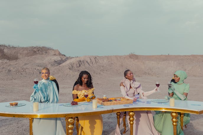 Elegant women in flowing gowns enjoying a glamorous sunset dinner in the desert dunes, surrounded by golden sand, lanterns, and laughter—blending adventure with luxury and feminine power.