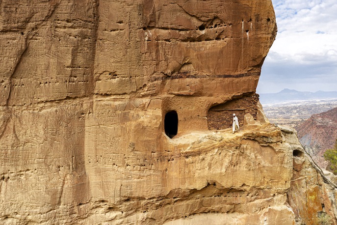 A remote rock-hewn church carved into a cliff face with a person walking along a narrow ledge, showcasing Abuna Yemata Guh in Ethiopia.