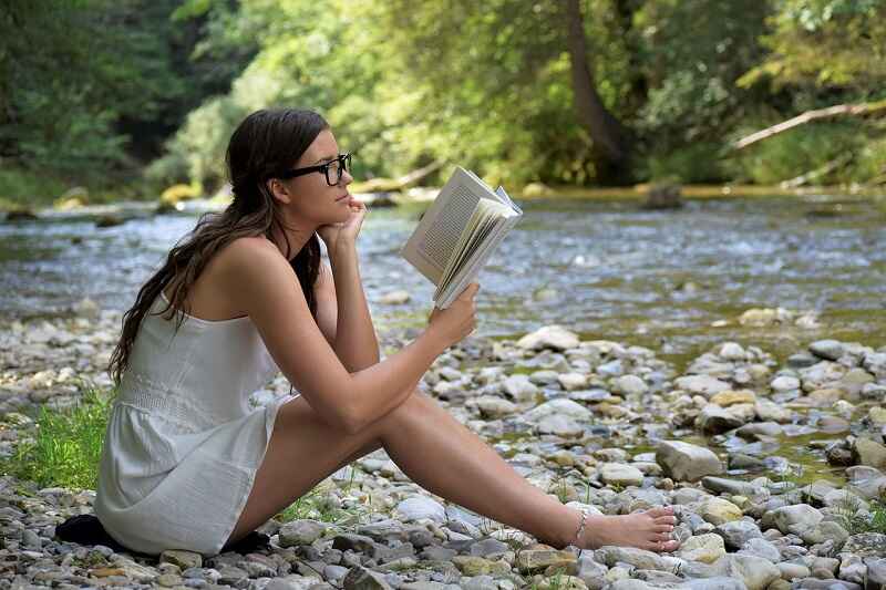A girl with spectacles sitting by a stream reading a book, relaxing in nature and enjoying wellness