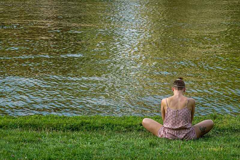 A woman sitting and relaxing on green grass by the side of a river.
